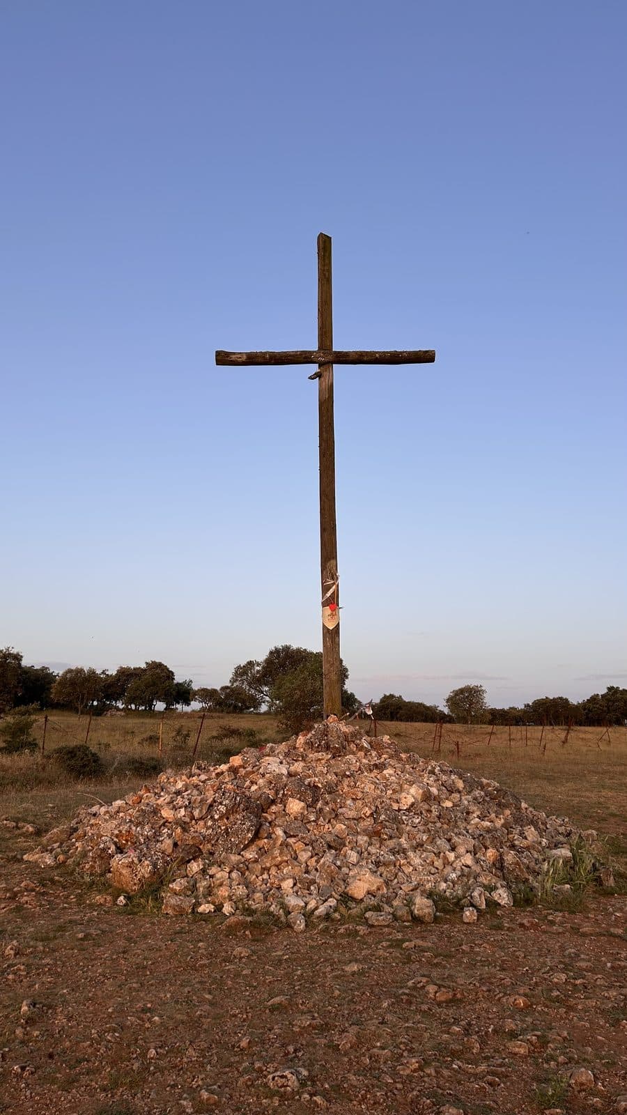 A plain wooden cross rising from a mound of pilgrim stones against a dusk sky.