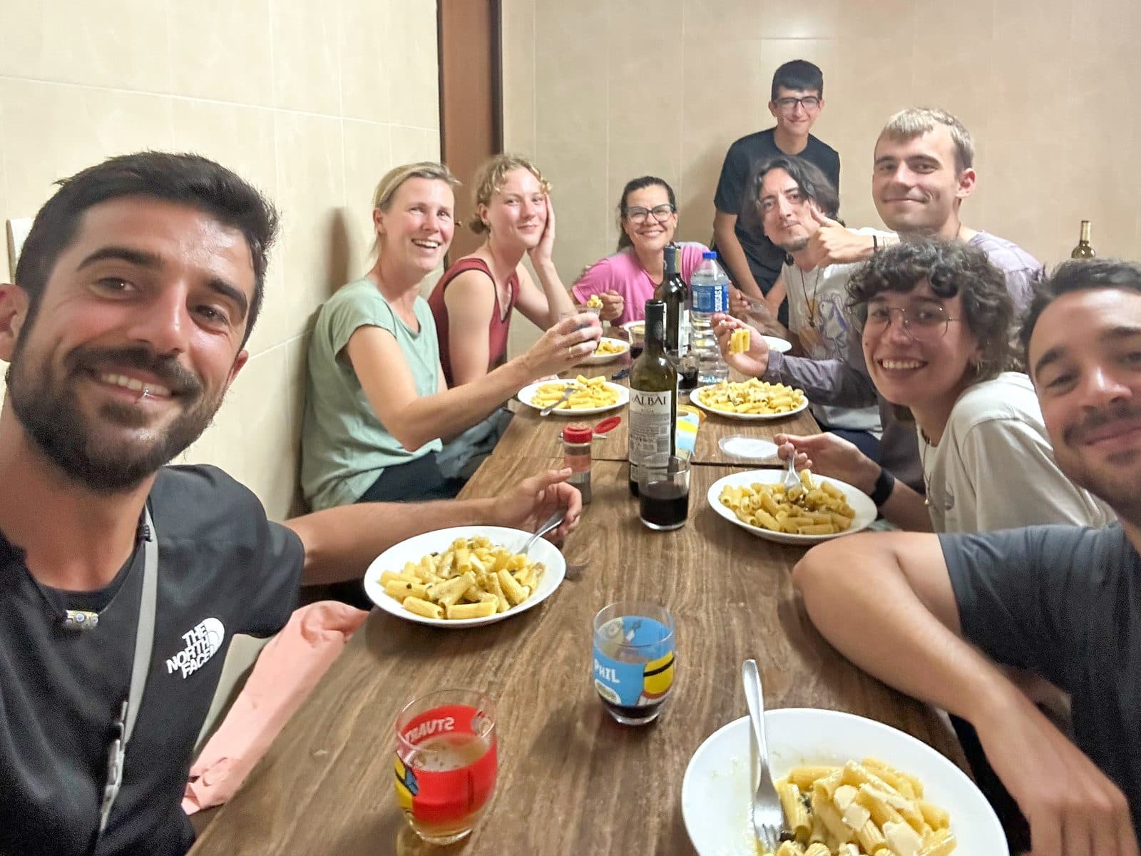 A communal pilgrim dinner around one table — pasta, wine, and laughter.