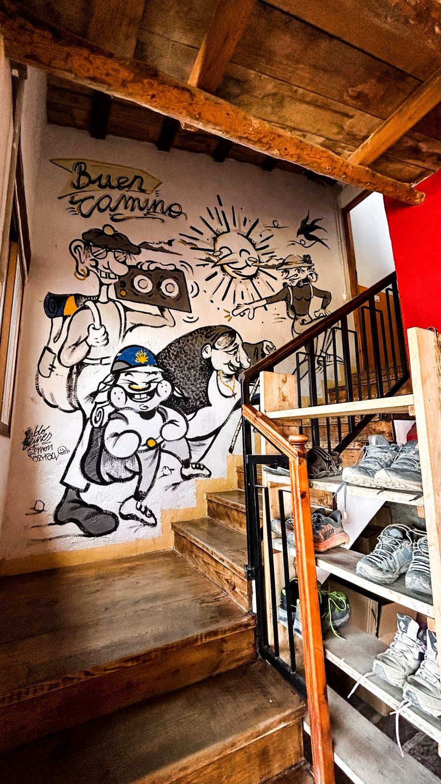 An albergue stairwell with hiking boots shelved below a hand-painted Buen Camino mural.