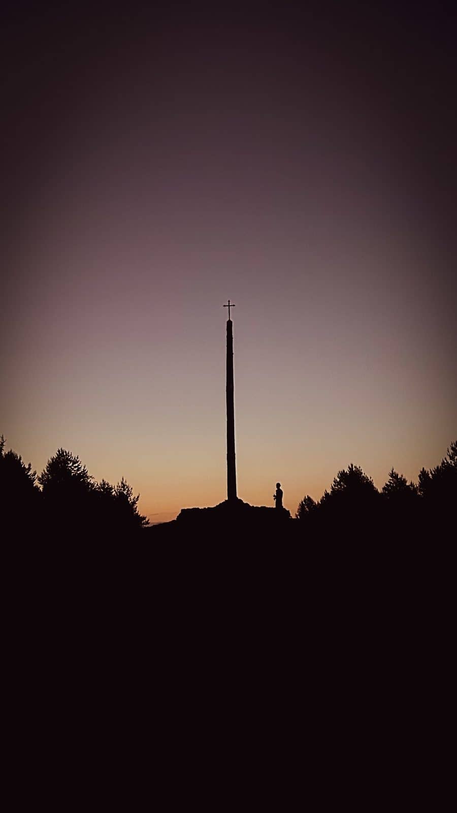 The silhouette of the Cruz de Ferro rising from its mound of pilgrim stones against a violet dawn sky.