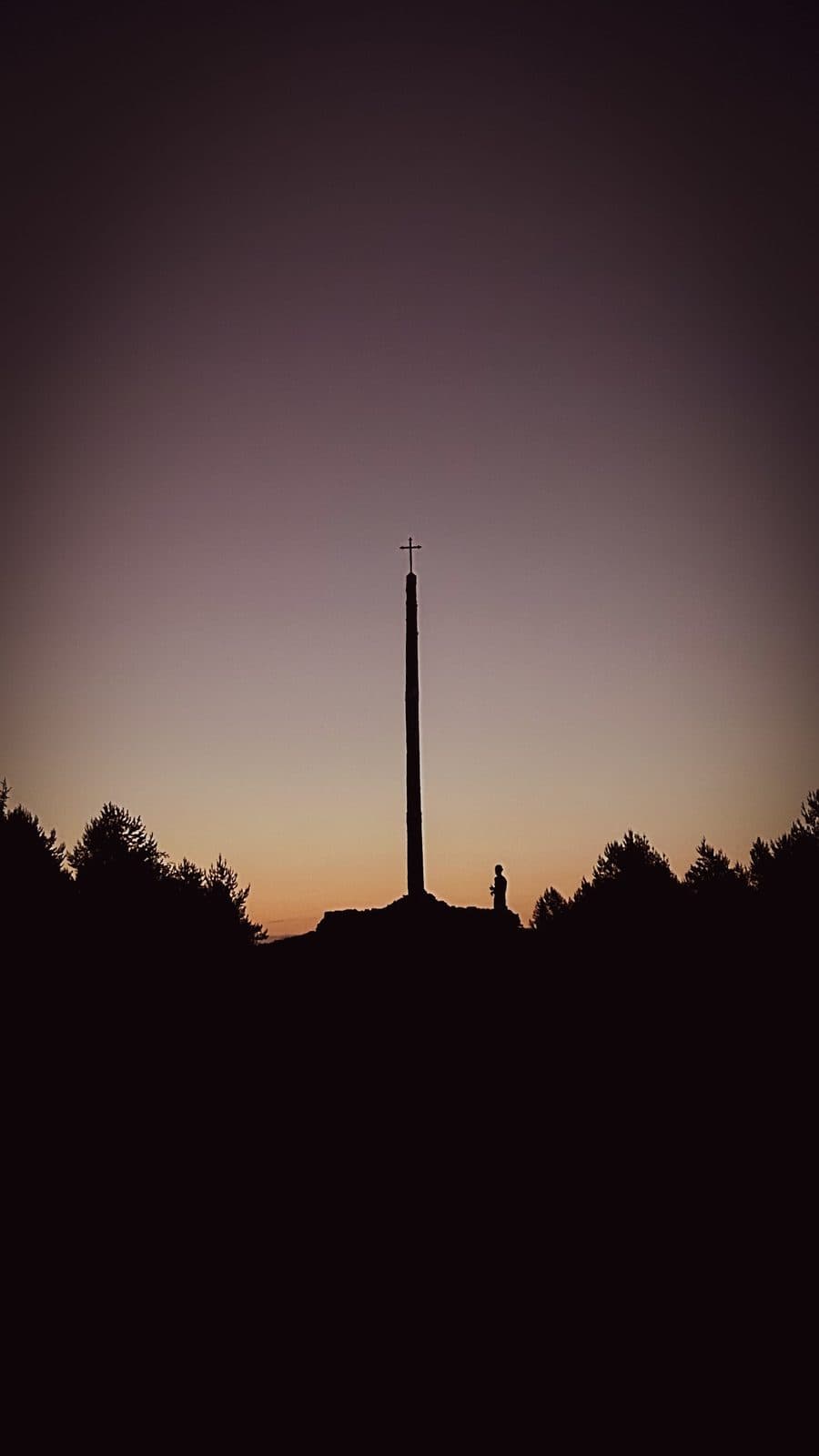 The silhouette of the Cruz de Ferro rising from its mound of pilgrim stones against a violet dawn sky.