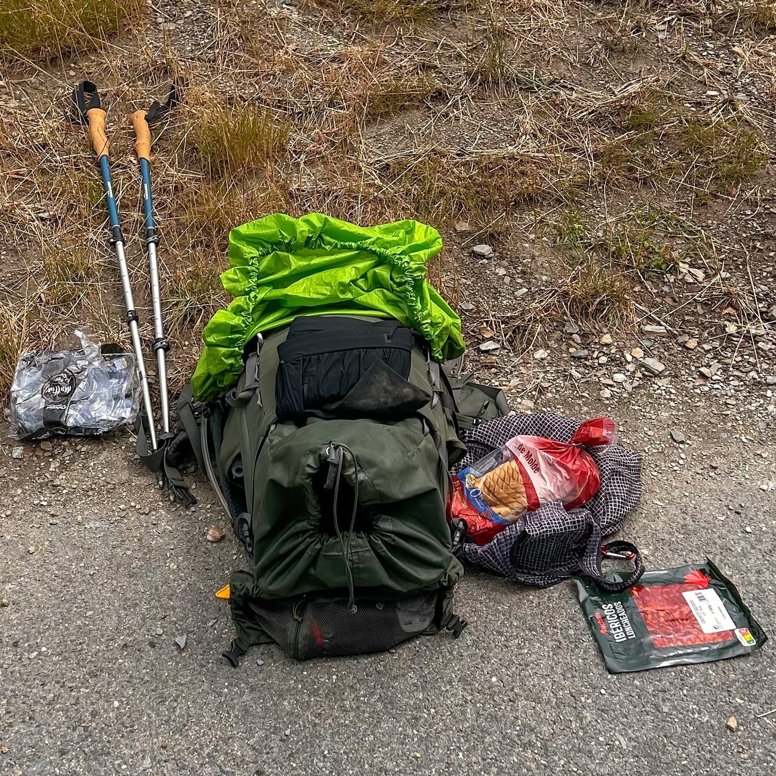 A loaded backpack, trekking poles, and the day's provisions laid out on gravel beside the trail.