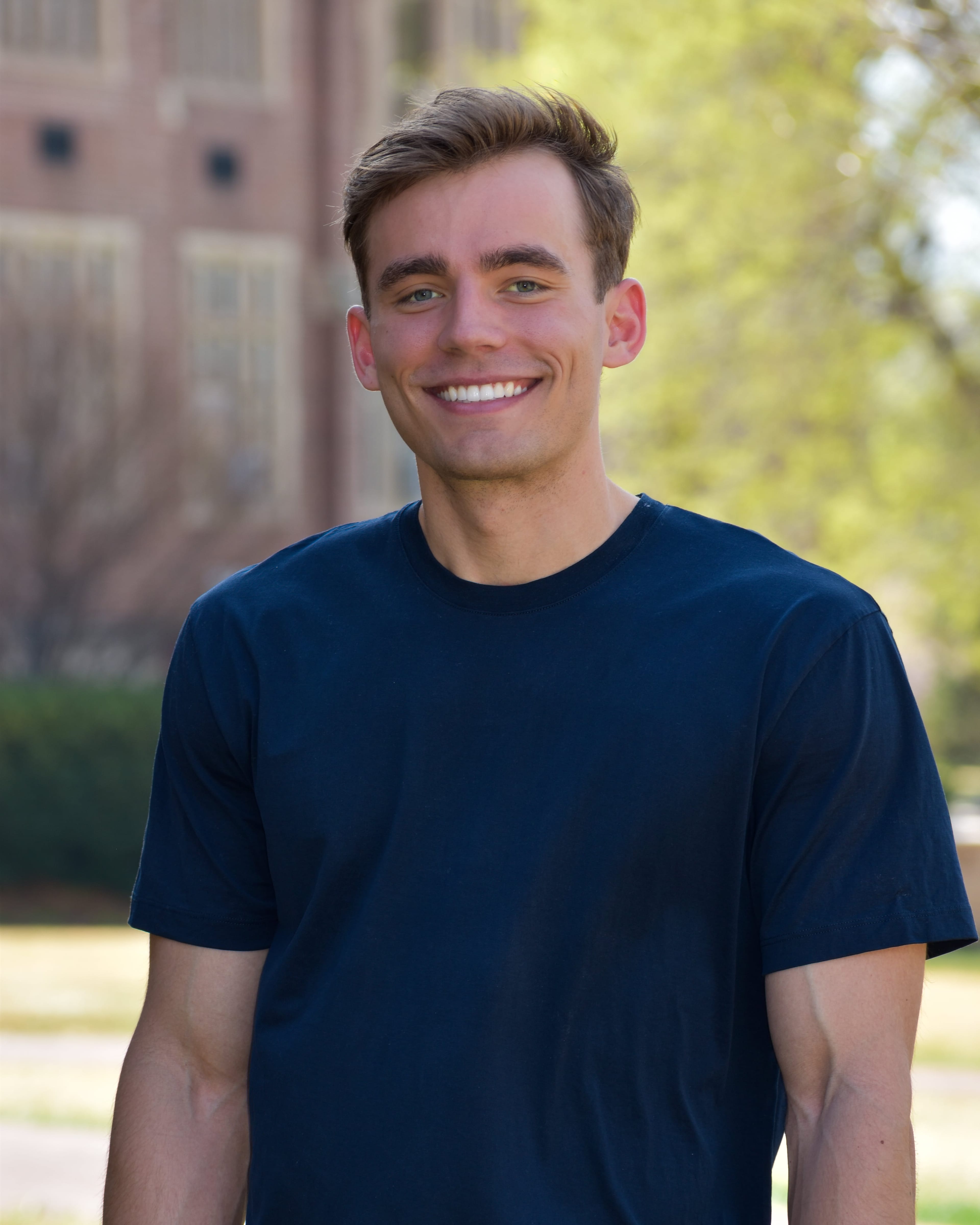 Kyle Wisniewski outdoors on a university campus, in a navy crew-neck shirt with a brick academic building and green trees behind him