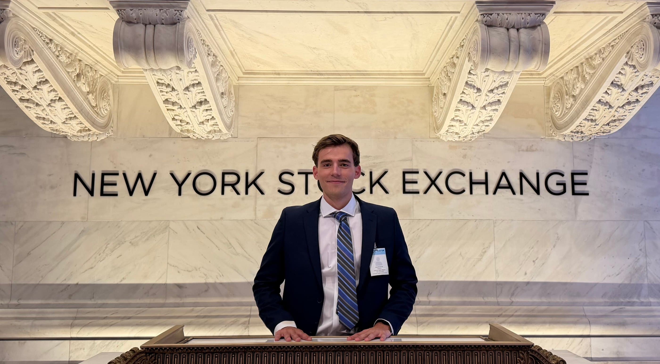 Kyle Wisniewski in a navy suit and striped tie, both hands on the marble bell podium of the New York Stock Exchange, the words NEW YORK STOCK EXCHANGE on the wall behind him