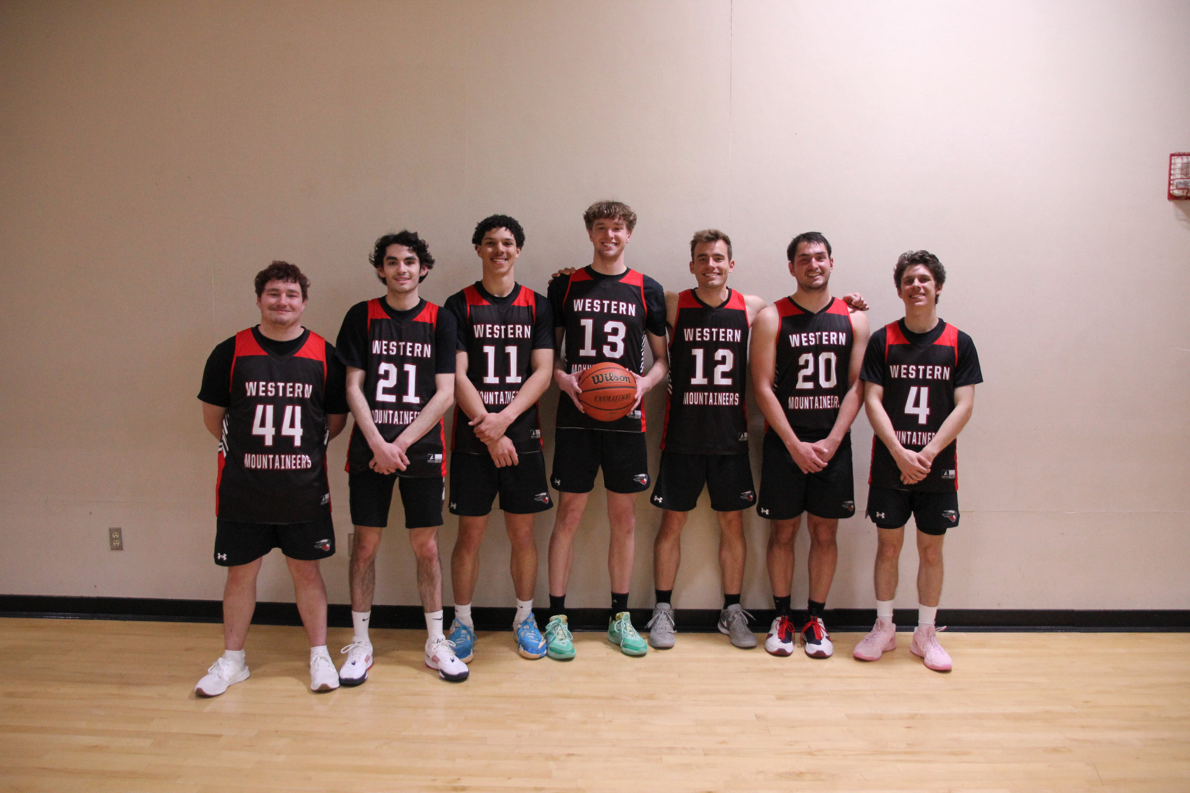 Seven players in black Western Mountaineers jerseys standing against a gym wall; Kyle Wisniewski wears 12.