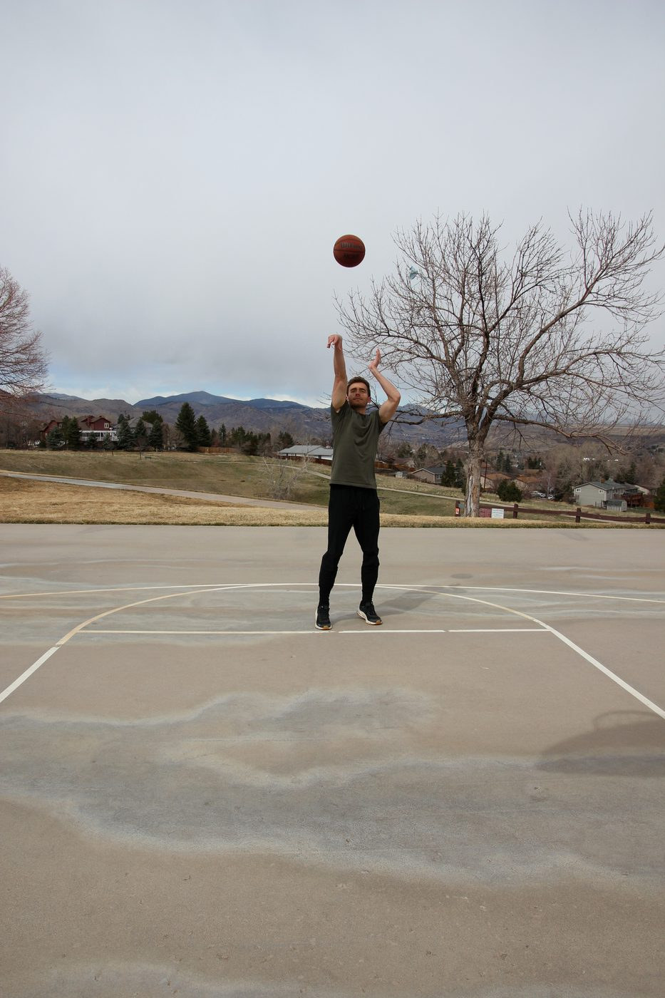 Kyle Wisniewski on an outdoor court following through on a jump shot, the ball high against a grey sky, foothills behind.