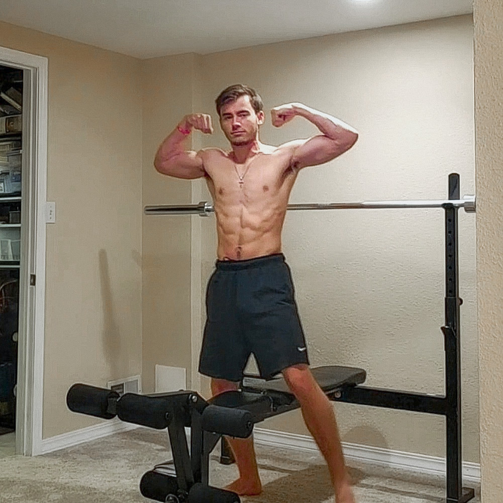 Kyle Wisniewski in dark shorts holding a front double-biceps pose beside a weight bench and barbell.