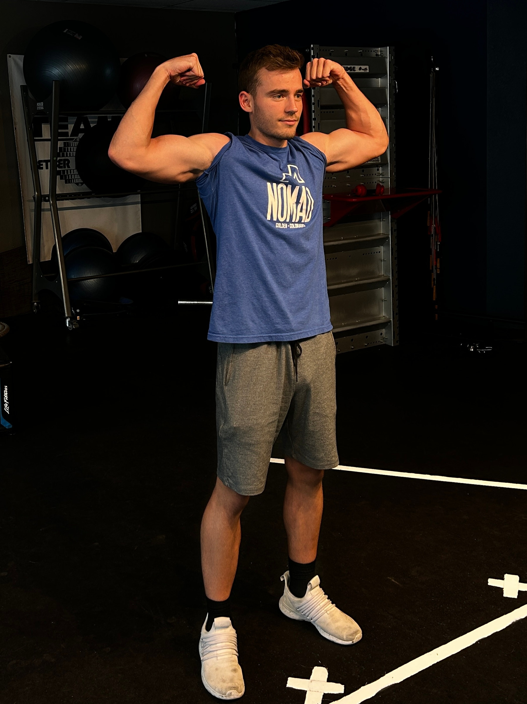 Kyle Wisniewski in a blue sleeveless shirt, grey shorts and white trainers, holding a front double-biceps pose in a training studio.
