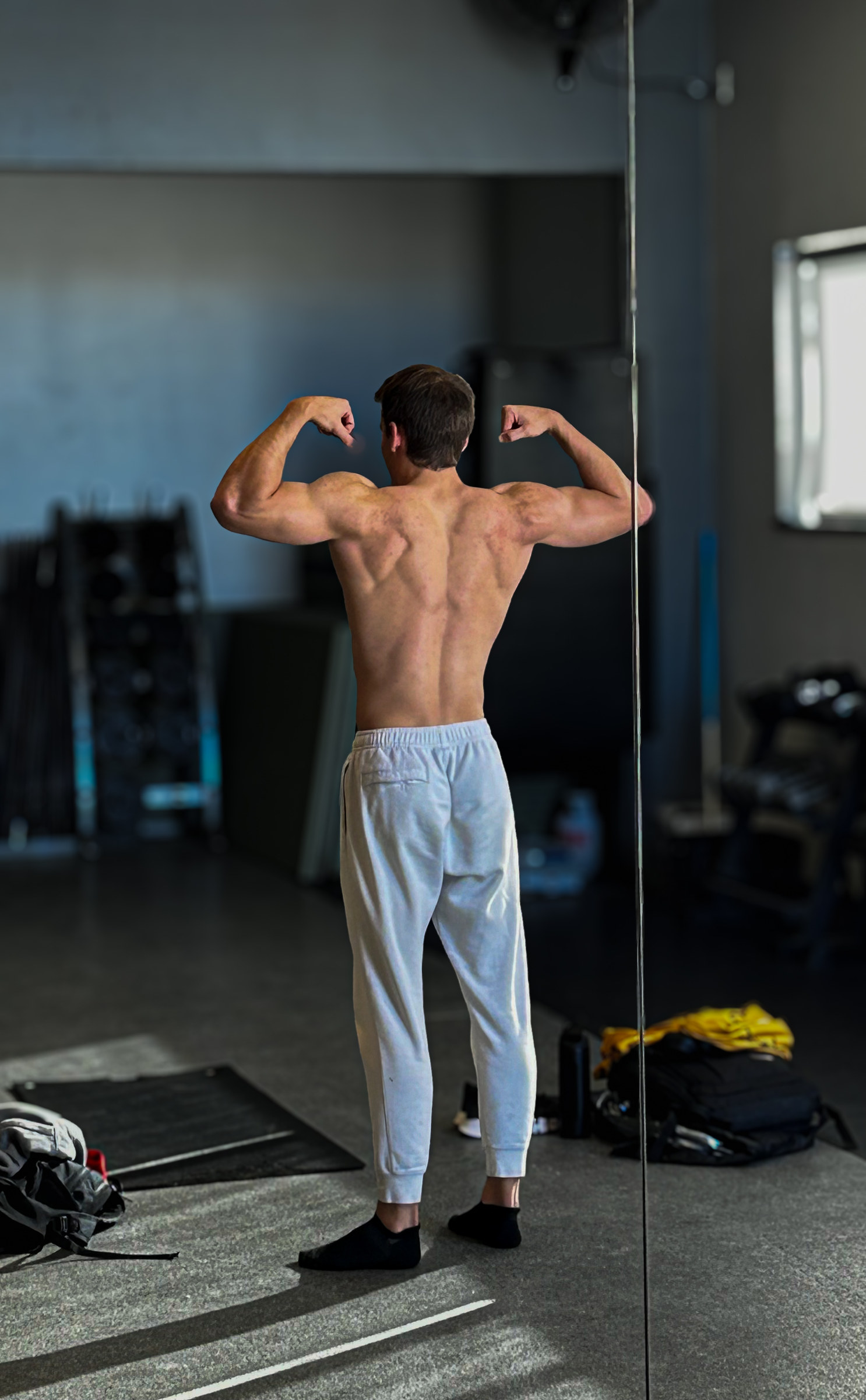 Kyle Wisniewski from behind in grey joggers, holding a back double-biceps pose near a window.