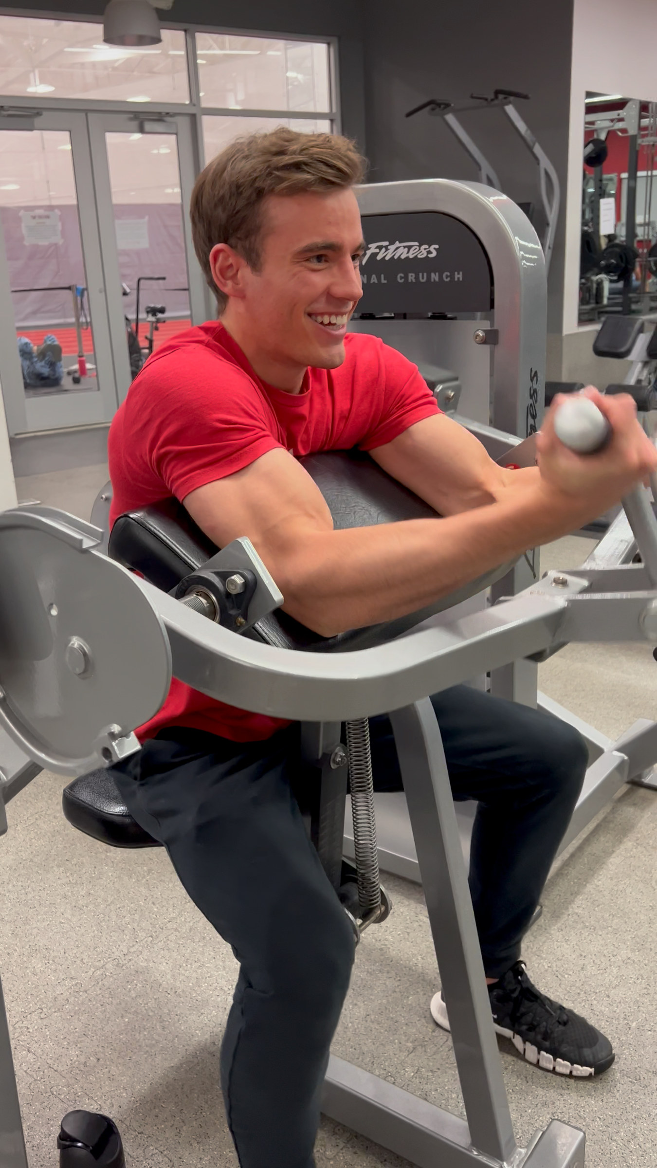 Kyle Wisniewski in a red T-shirt, smiling, mid-repetition on a preacher-curl machine.