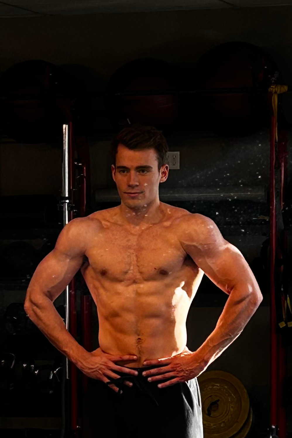 Kyle Wisniewski with hands on hips, lit by low window light in a dark gym with racks and plates.