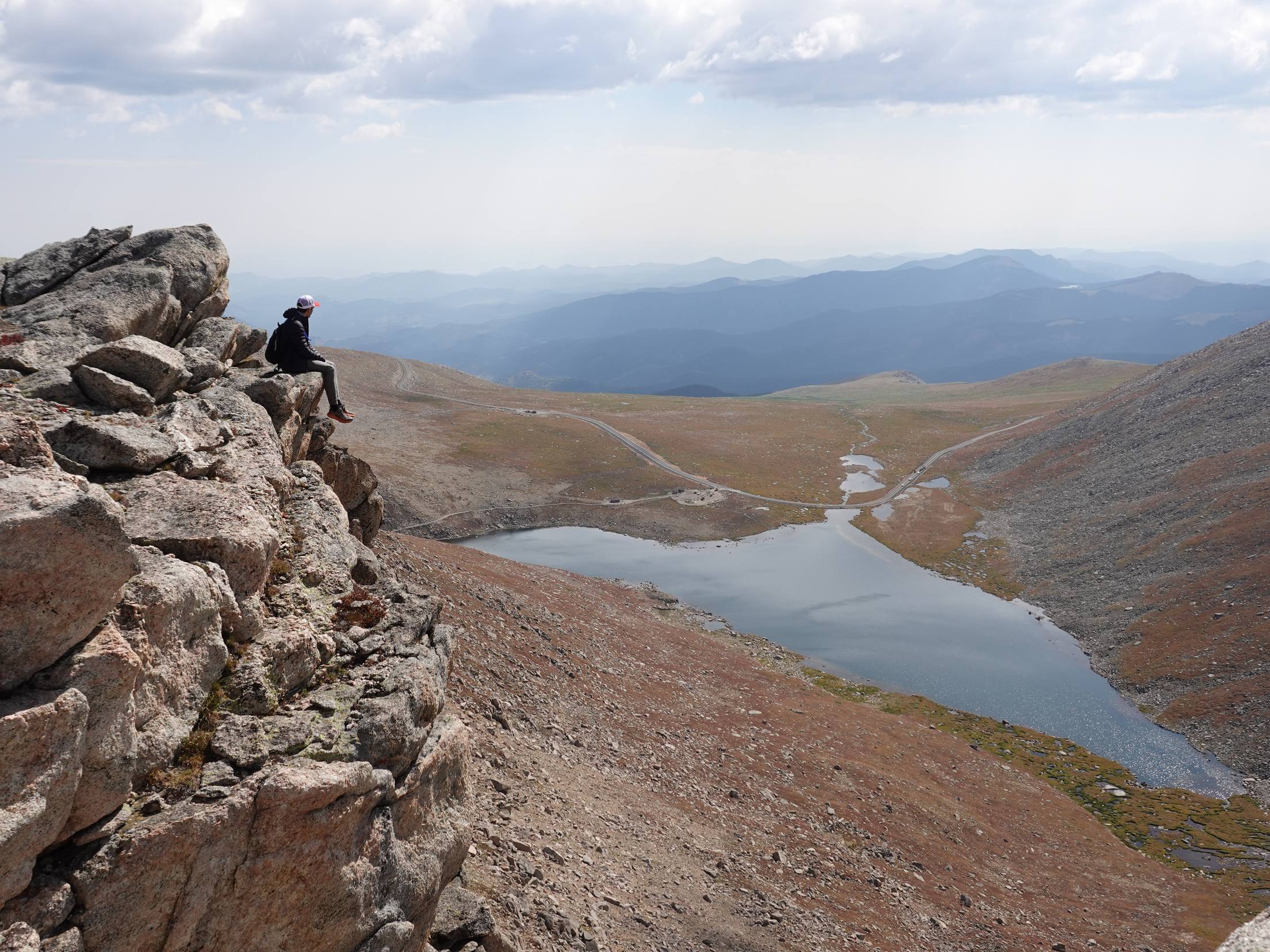 Kyle Wisniewski seated on a granite outcrop high above a lake, a winding road in the basin below and blue mountain ranges beyond.