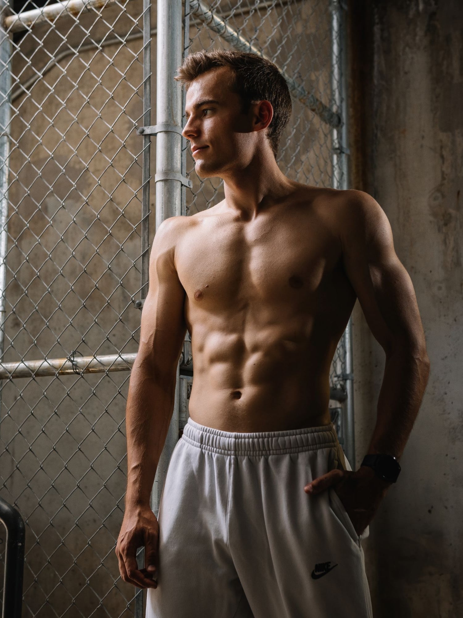 Kyle Wisniewski, shirtless in grey sweatpants, one hand in a pocket, looking aside by a chain-link fence and concrete wall