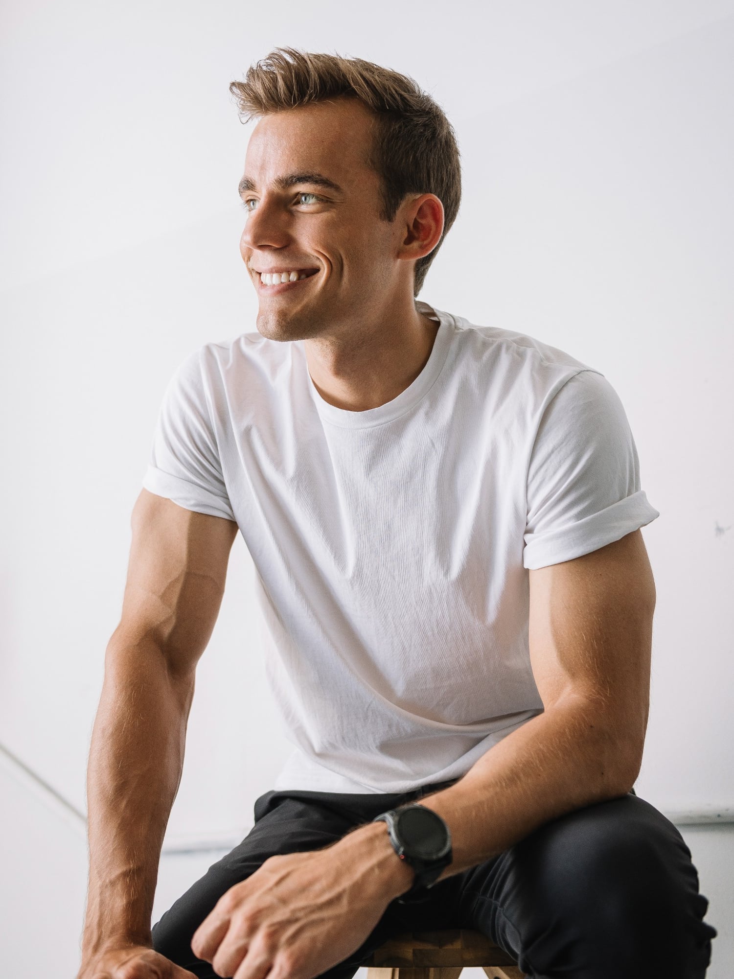 Kyle Wisniewski, seated in a white T-shirt and black trousers, leaning forward, smiling and looking to one side