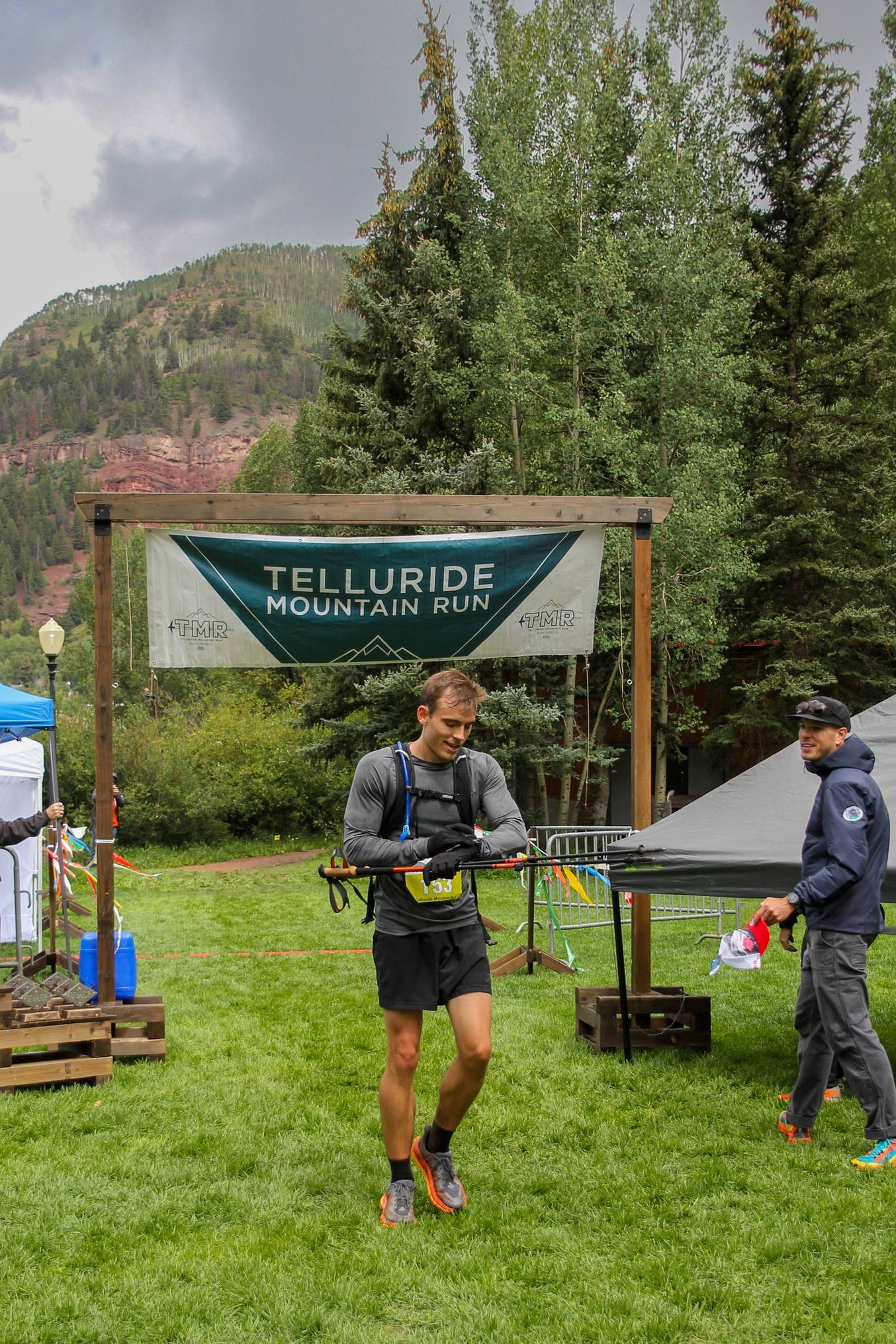 Kyle Wisniewski in a grey shirt with race vest and poles, wearing bib 153, walking on grass beneath the Telluride Mountain Run banner.