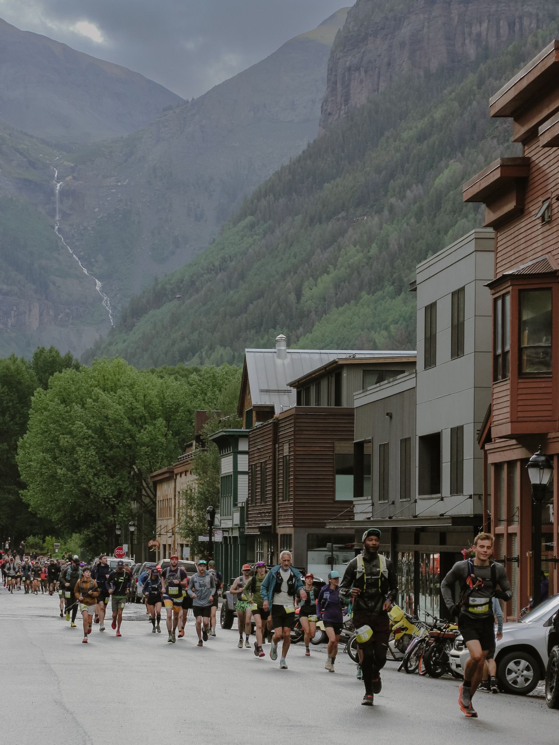 Runners in race vests on a Telluride street, a waterfall and steep green mountains behind; Kyle Wisniewski at front right.