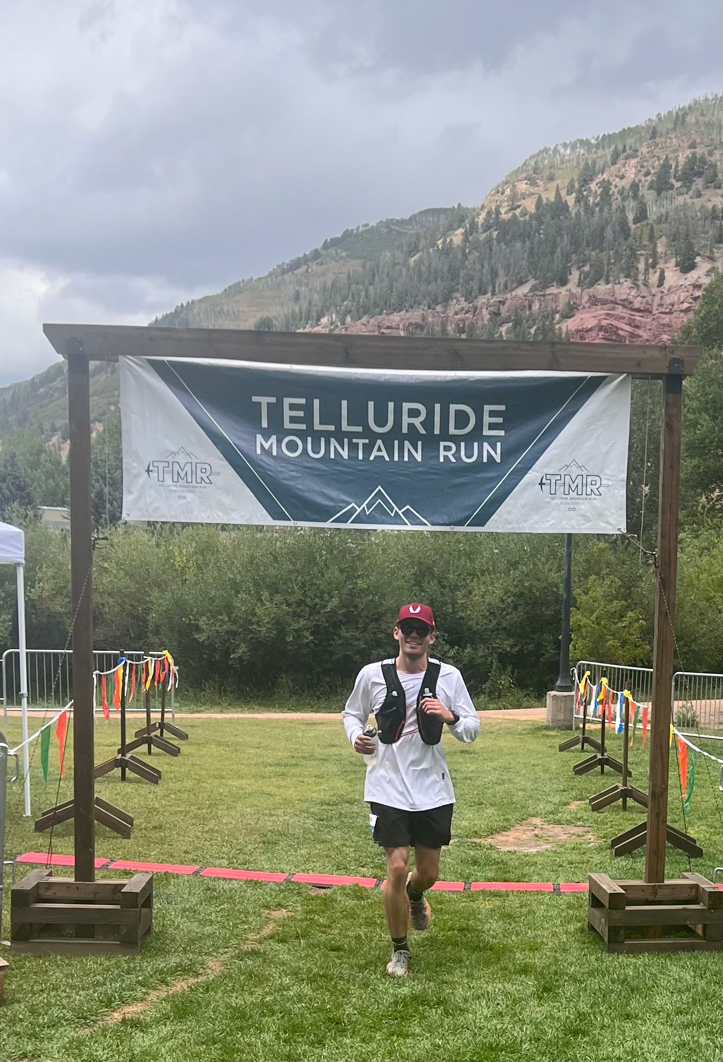 Kyle Wisniewski in a red cap, white long-sleeve shirt and race vest, running toward the camera under the Telluride Mountain Run banner on a grass field.