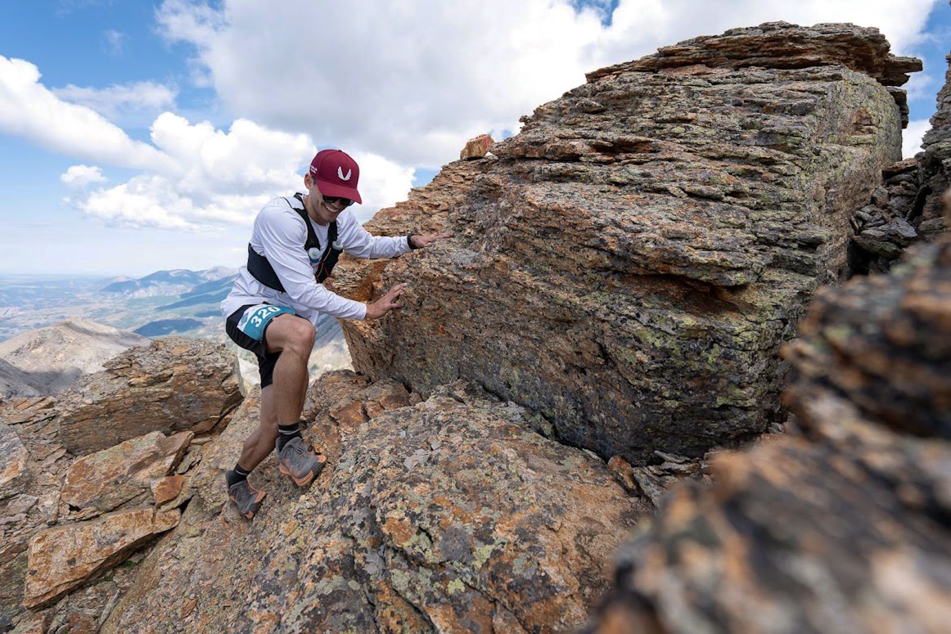 Kyle Wisniewski in a red cap, white long-sleeve shirt and race vest, climbing a lichen-covered rock outcrop with mountain ranges behind him.