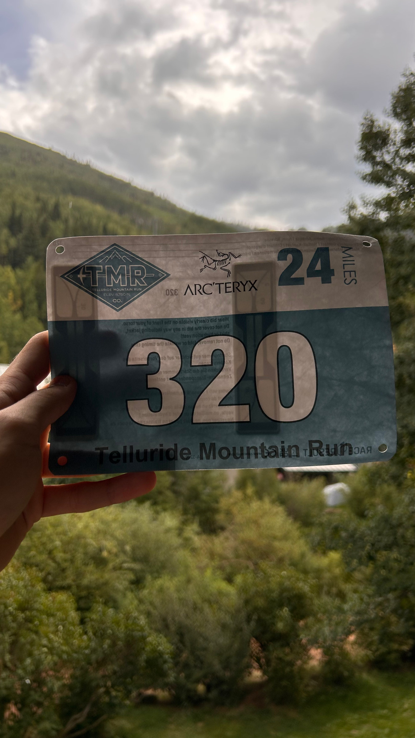 A hand holding race bib 320, Telluride Mountain Run 24 Miles, against a green hillside.