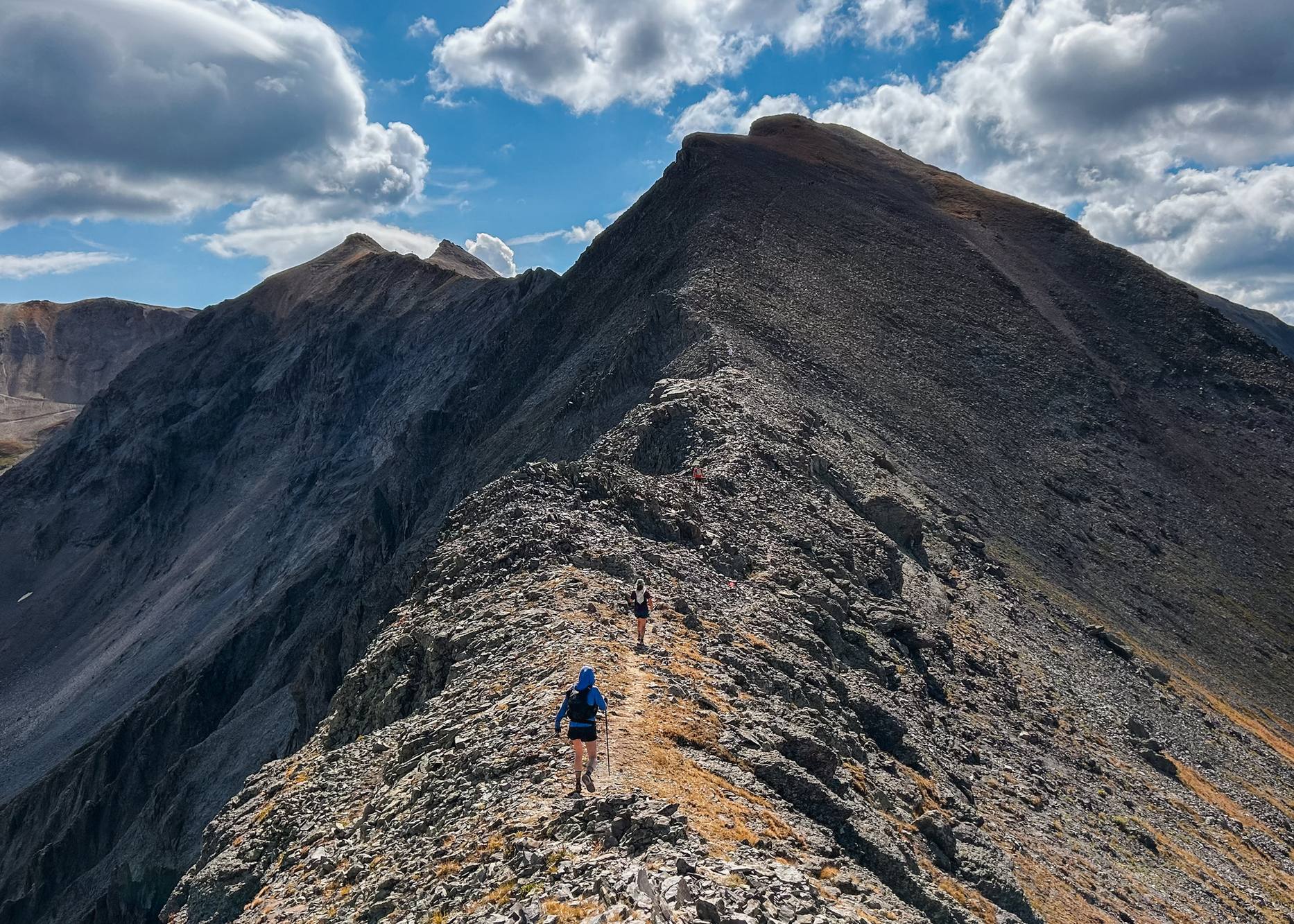 Two runners on a narrow rocky ridge beneath a dark summit, blue sky and white clouds above.