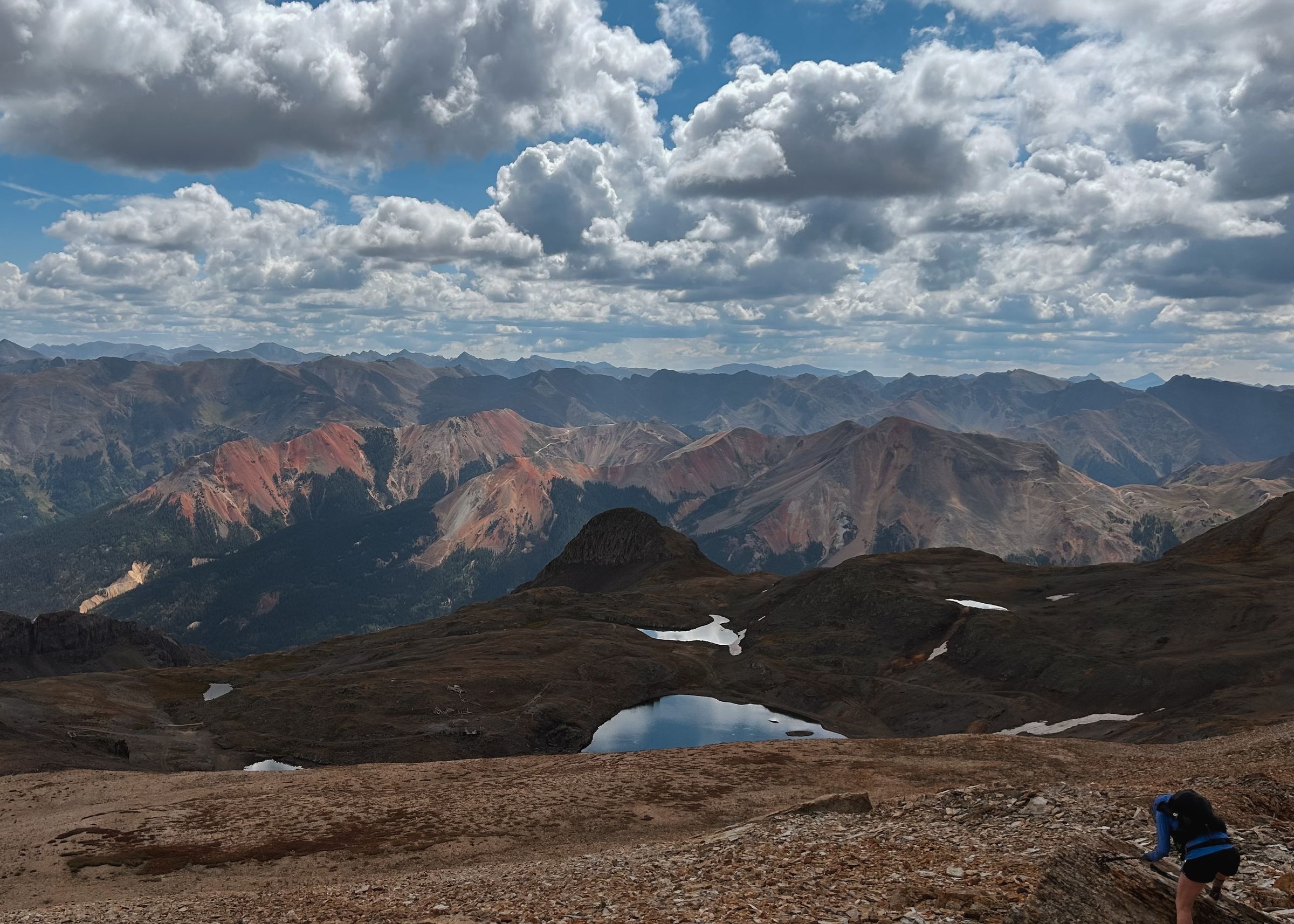 Two small alpine lakes in a high basin below red and grey peaks under scattered cloud; a runner at the lower right edge.