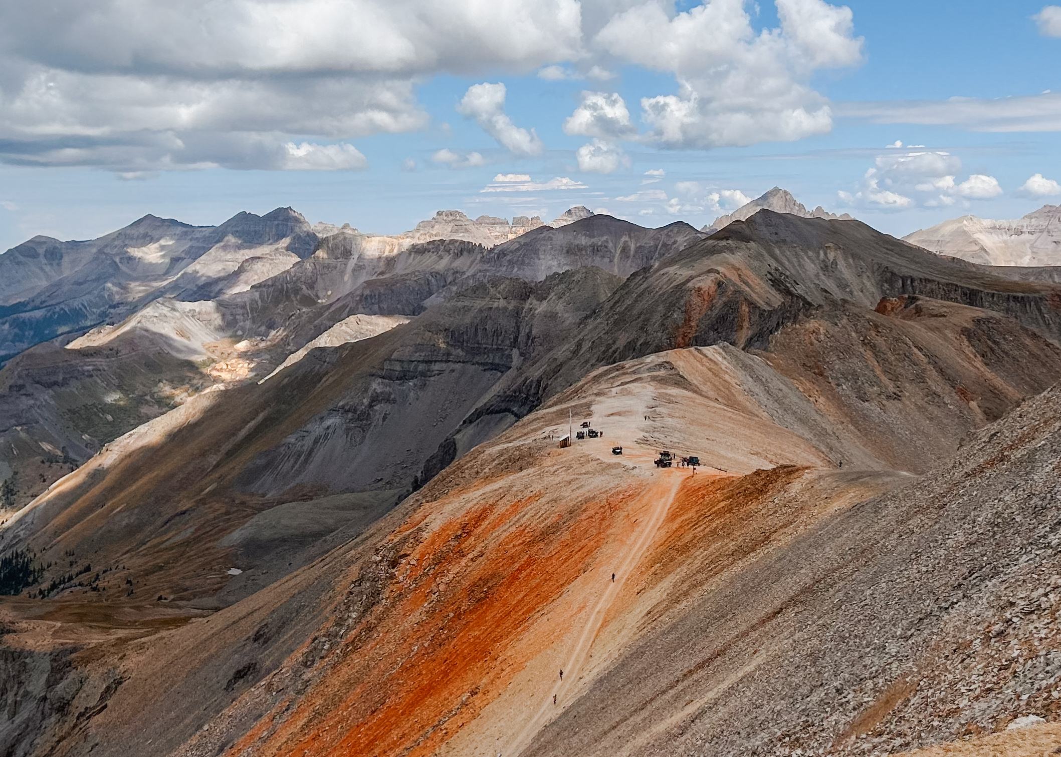 A trail crossing red and orange scree beneath grey peaks, small figures on the ridge.