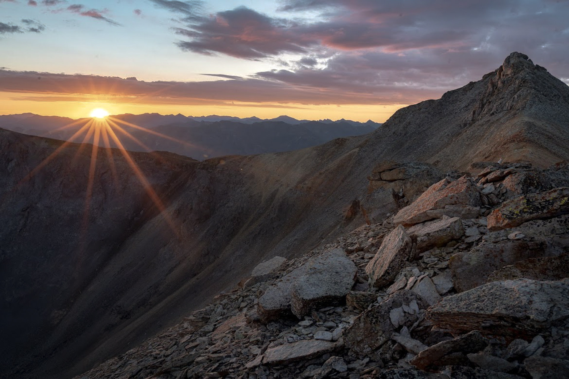 The sun low over a dark mountain ridge, rays across a rocky slope.